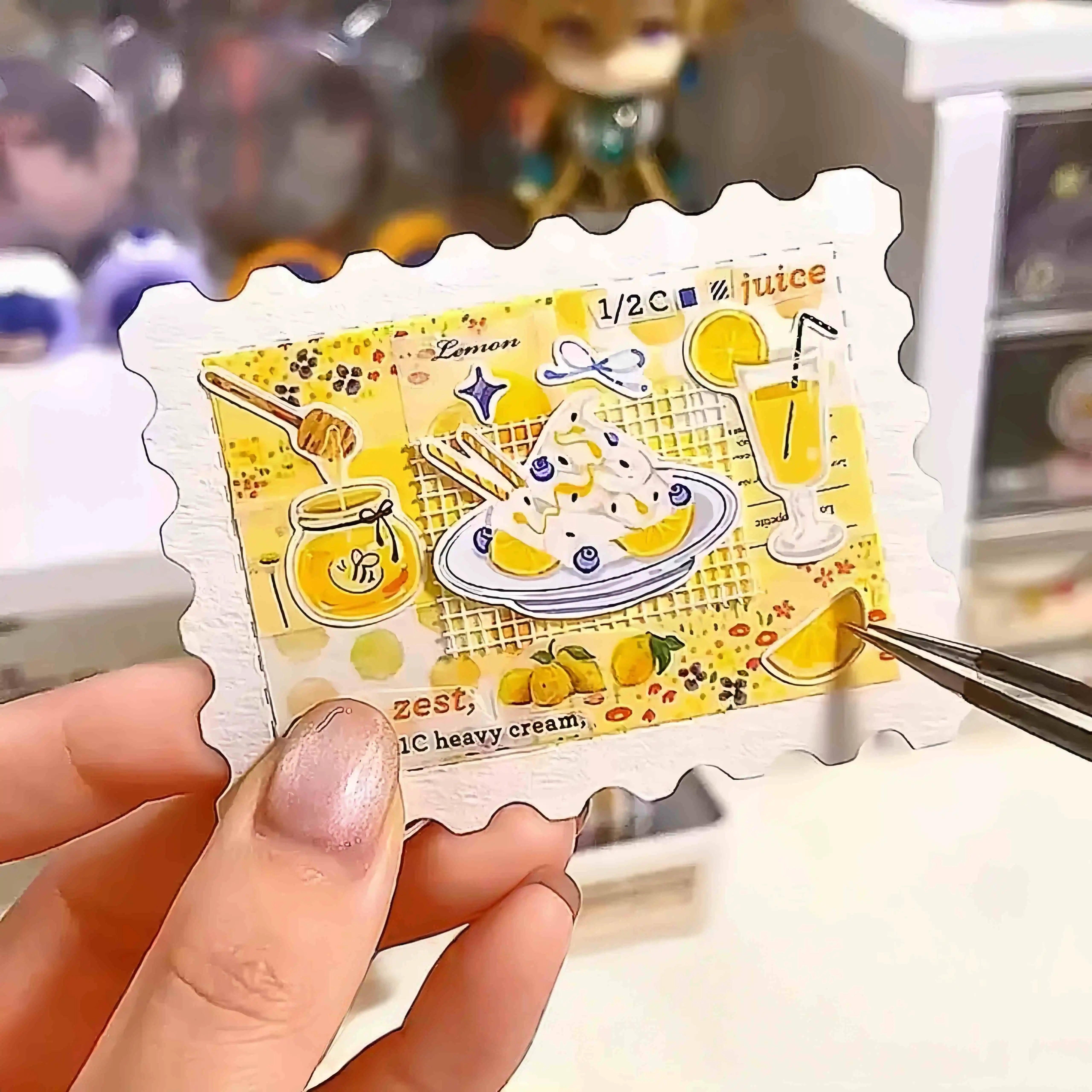 Completed journal insert with colorful food illustration painted on a blank stamp-shaped watercolor card, held with tweezers