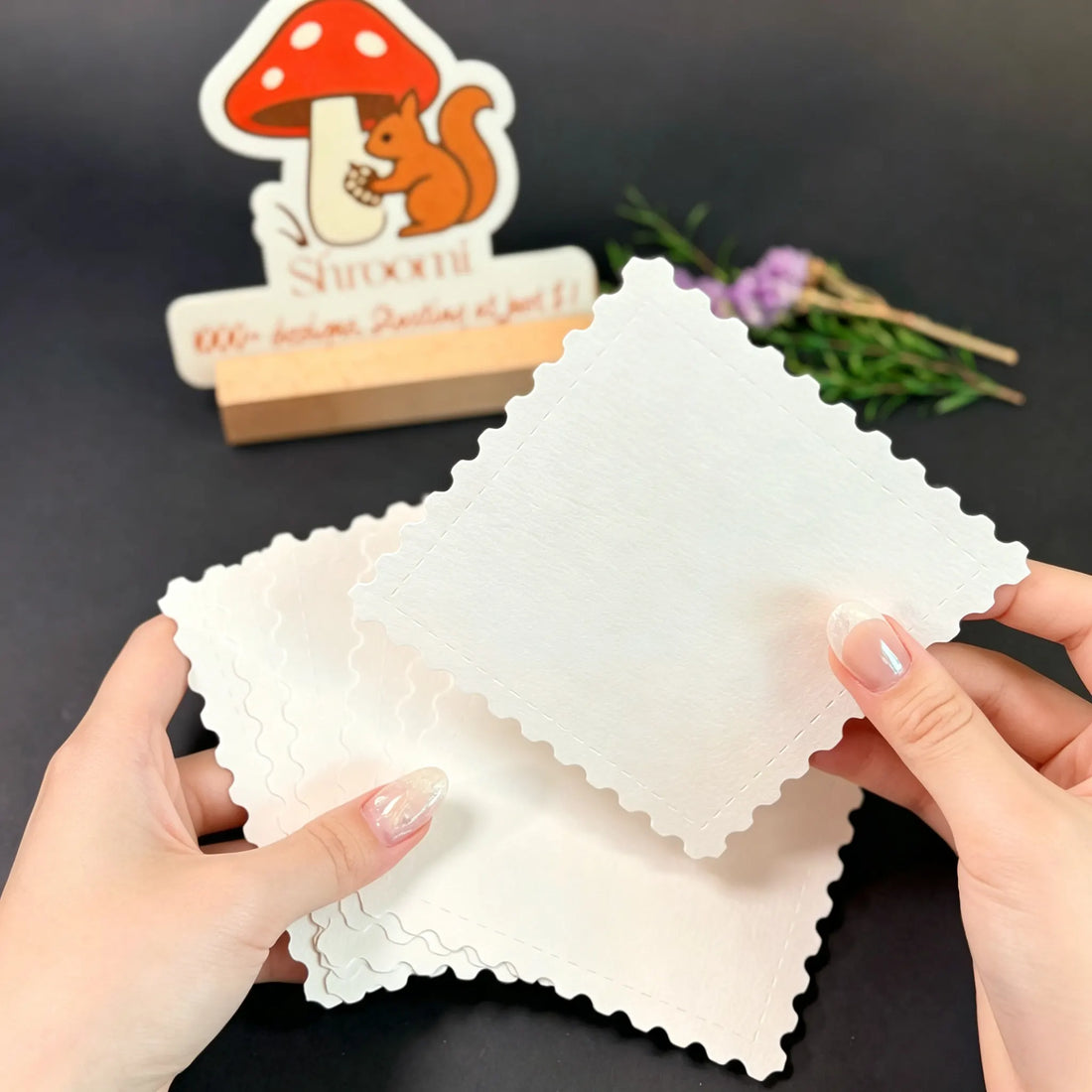 Hands holding a single 5.5x5.5cm blank square stamp-shaped watercolor paper card with scalloped edges