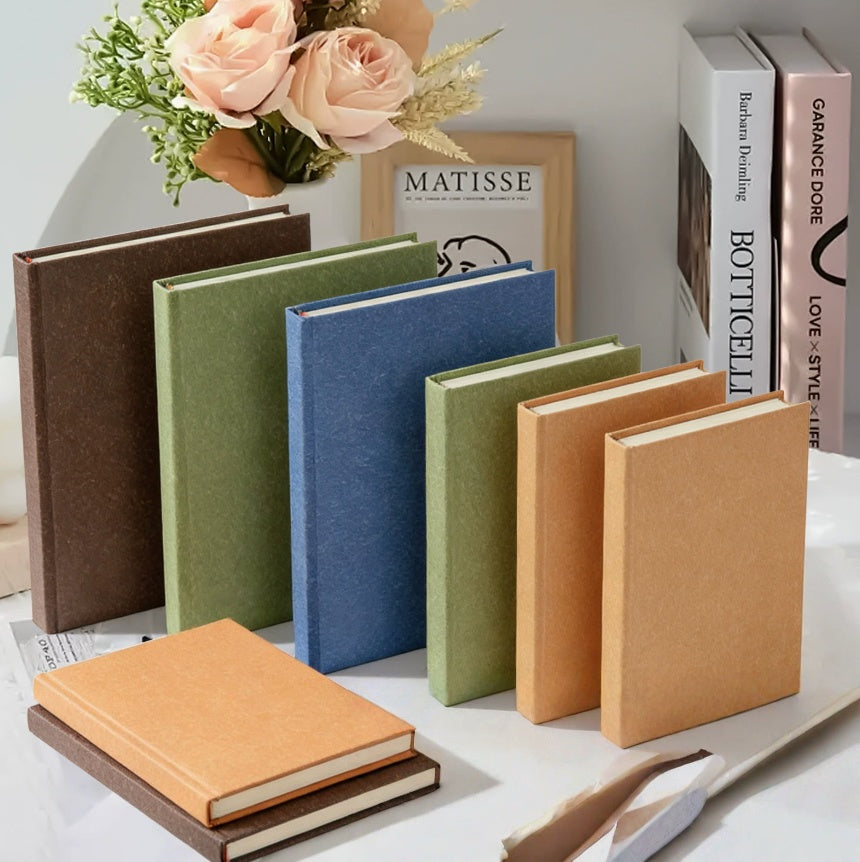 Four A6 hardcover blank journals in brown, olive, blue and orange natural texture covers on grass with colored pencils