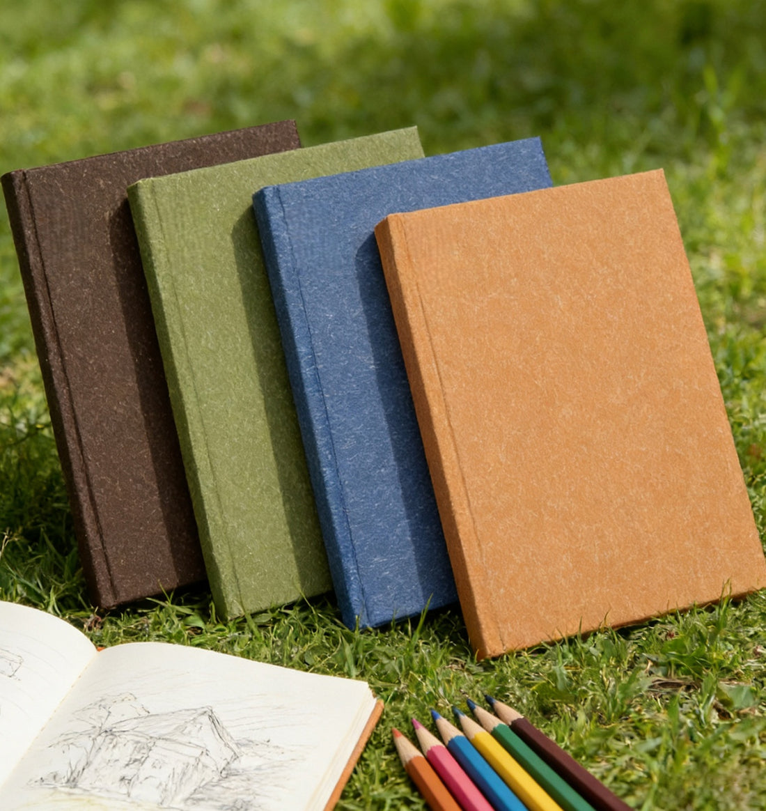 Four A6 hardcover blank journals in brown, olive, blue and orange natural texture covers on grass with colored pencils