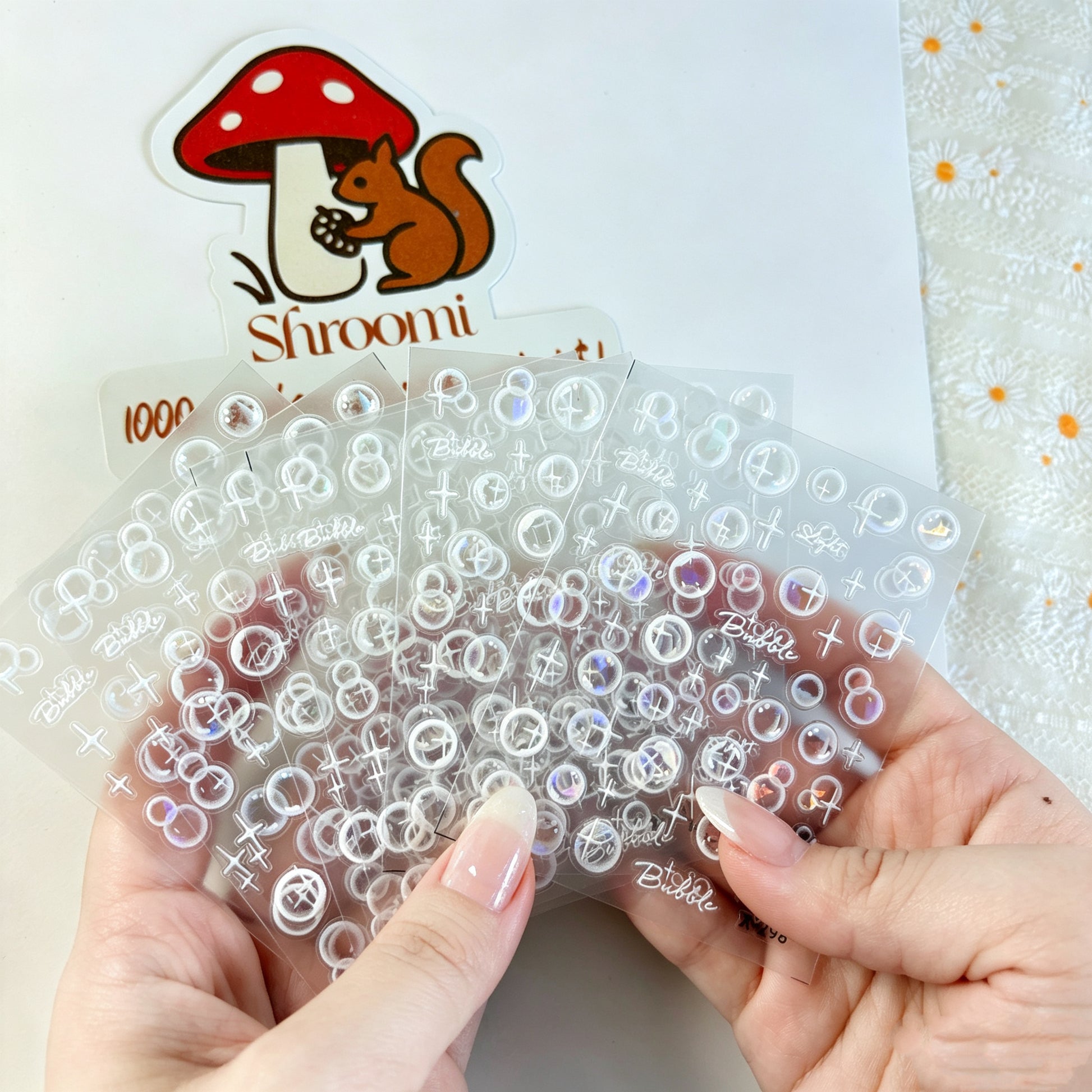 Hand holding five clear bubble sticker sheets with shimmer accents and Shroomi packaging in the background