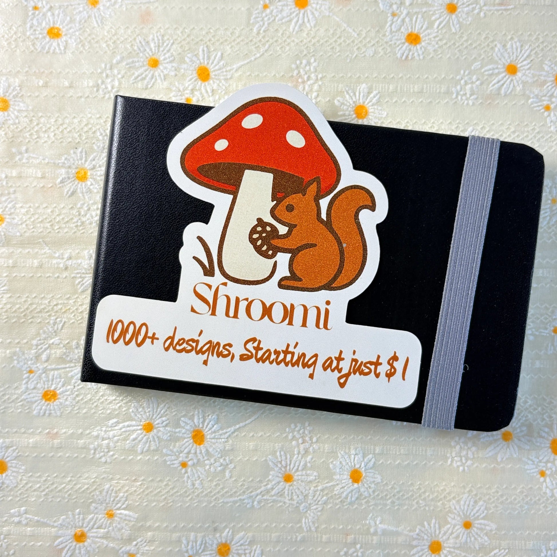 Black hardcover journal notebook laying flat showing Shroomi mushroom and squirrel sticker card