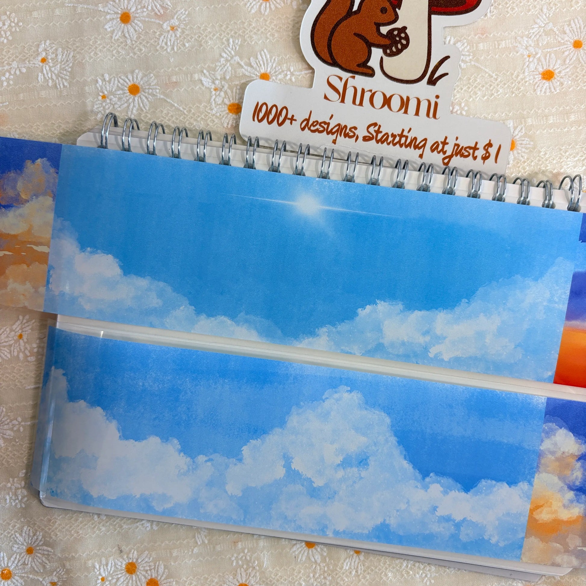 Shroomi Four Seasons Sky Background PET tape on journal — two strips: soft pale blue daytime sky with white fluffy clouds (top), warm orange sunset sky framed by deep blue-purple storm clouds (bottom)