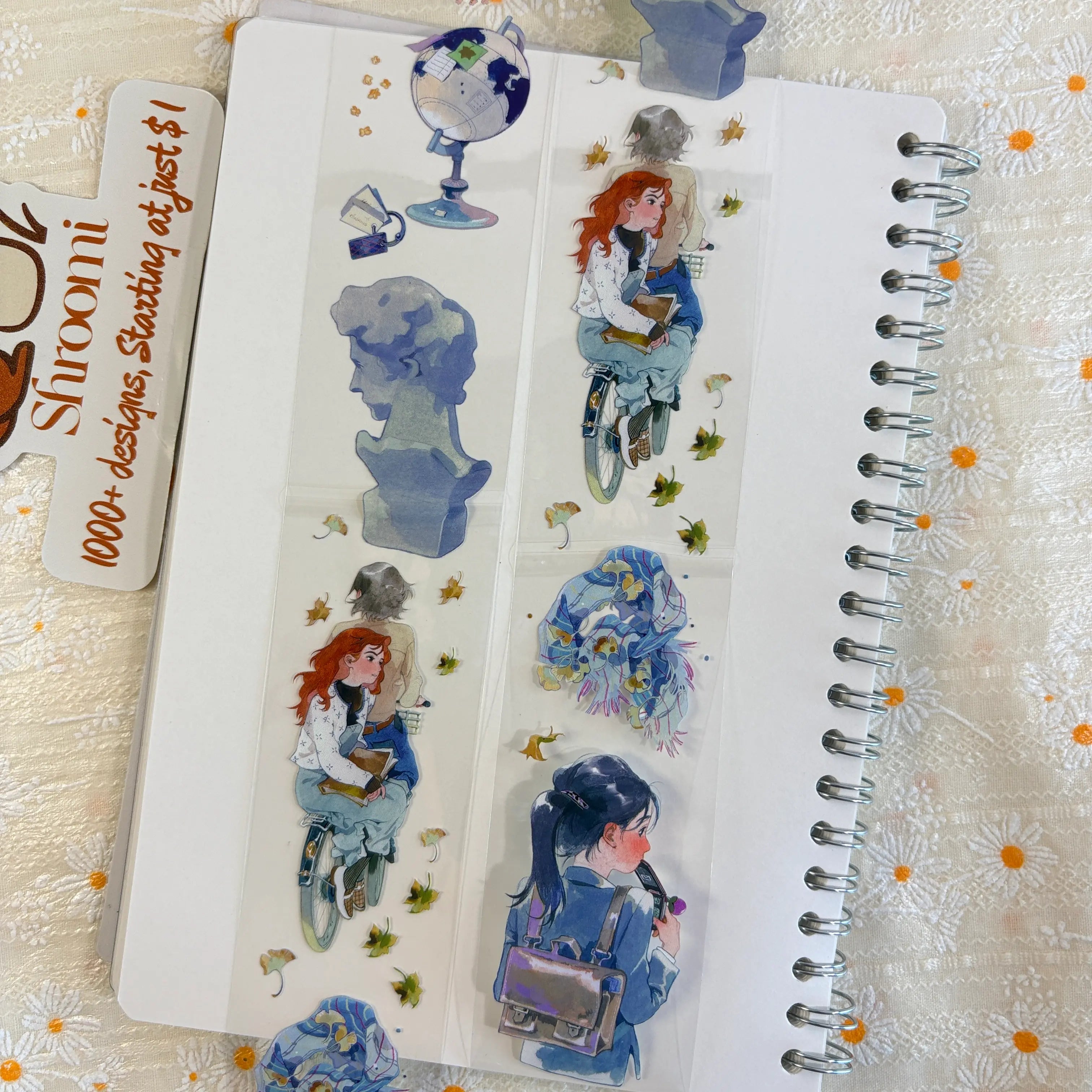 Shroomi PET tape on journal showing autumn campus scenes: globe on stand, classical grey bust sculpture, red-haired girl cycling with books and autumn leaves, girl with blue wildflowers and satchel backpack, falling ginkgo leaves