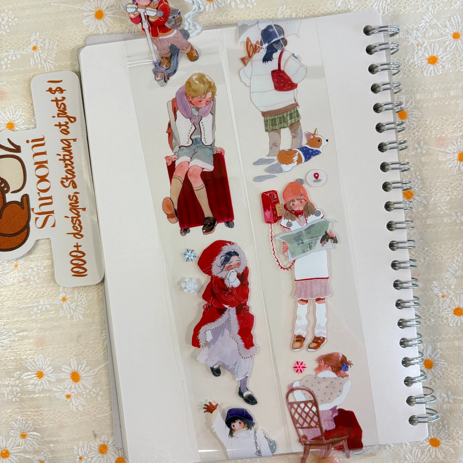 Shroomi PET tape on journal showing winter scenes: child sitting on red suitcase with purple scarf, girl in red parka with snowflakes, girl in orange hat reading a map with location pin, girl sitting in chair
