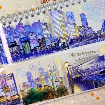 Shroomi Night City Shimmer PET tape on journal — two strips: blue-purple night skyline with illuminated skyscrapers and fireworks (top); city skyline with communications tower, fireworks, and lamp-lit bridge over water (bottom)